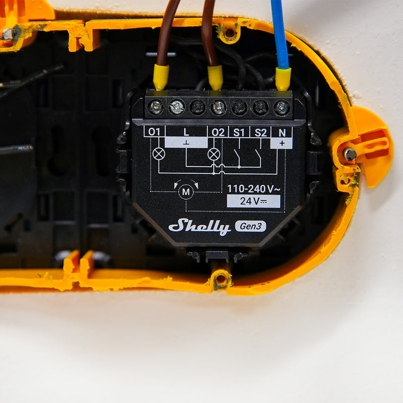 Close-up of a Shelly 2PM Gen3 x2 device installed in a yellow wall-mounted enclosure, showing wiring connections and a front panel diagram.